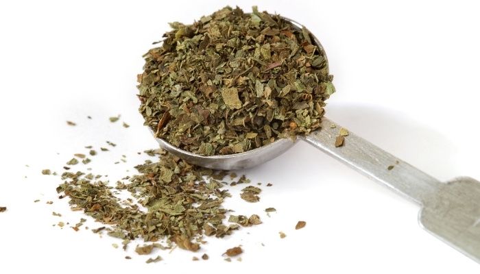 a metal scoop filled with green tea next to a pile of dried herbs on a white surface