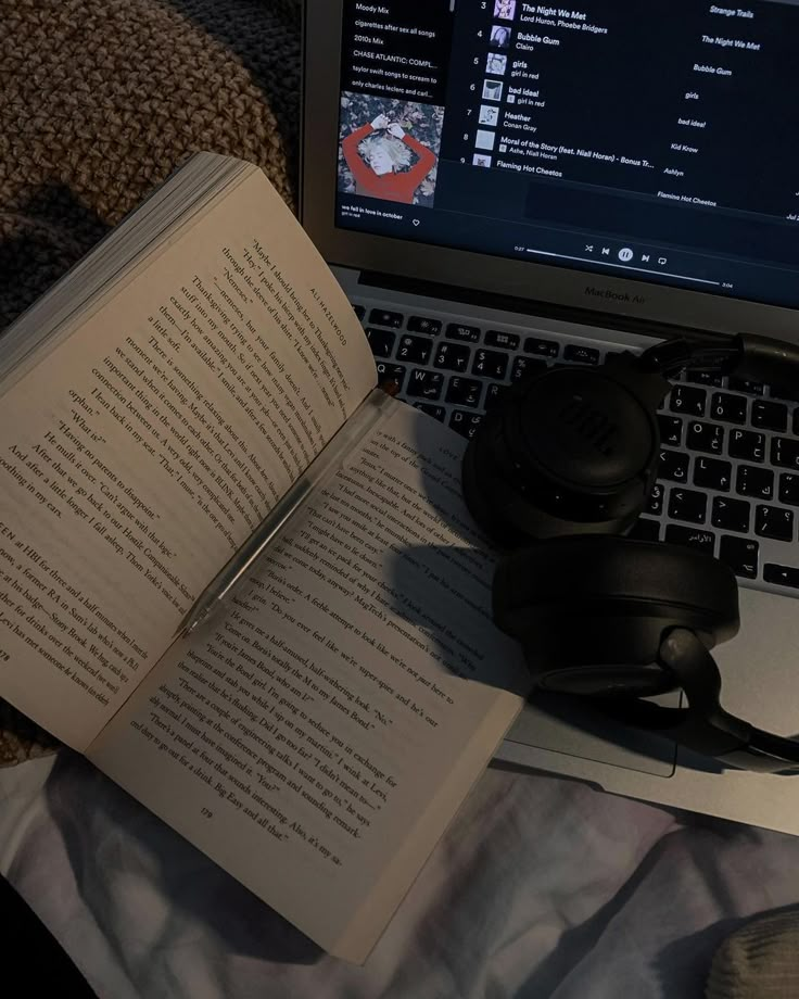 an open book sitting on top of a laptop computer next to headphones and books