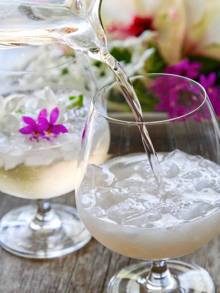 a pitcher pouring water into a glass filled with ice and flowers in the back ground