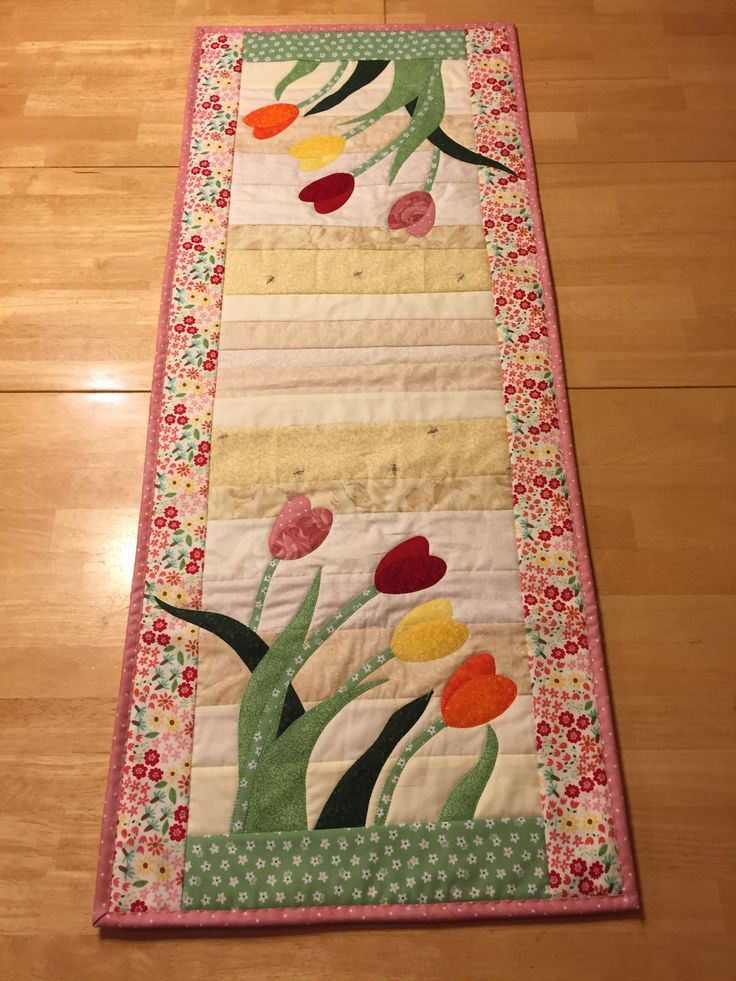 a quilted table runner with flowers and leaves on the border, sitting on a wooden floor