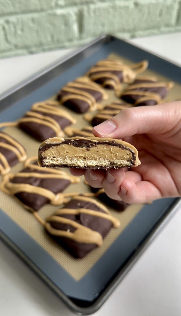 a hand holding a peanut butter cookie with chocolate drizzled on it, in front of a baking sheet