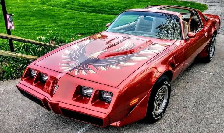 a red pontiac firebird parked in front of a wooden fence and green grass behind it