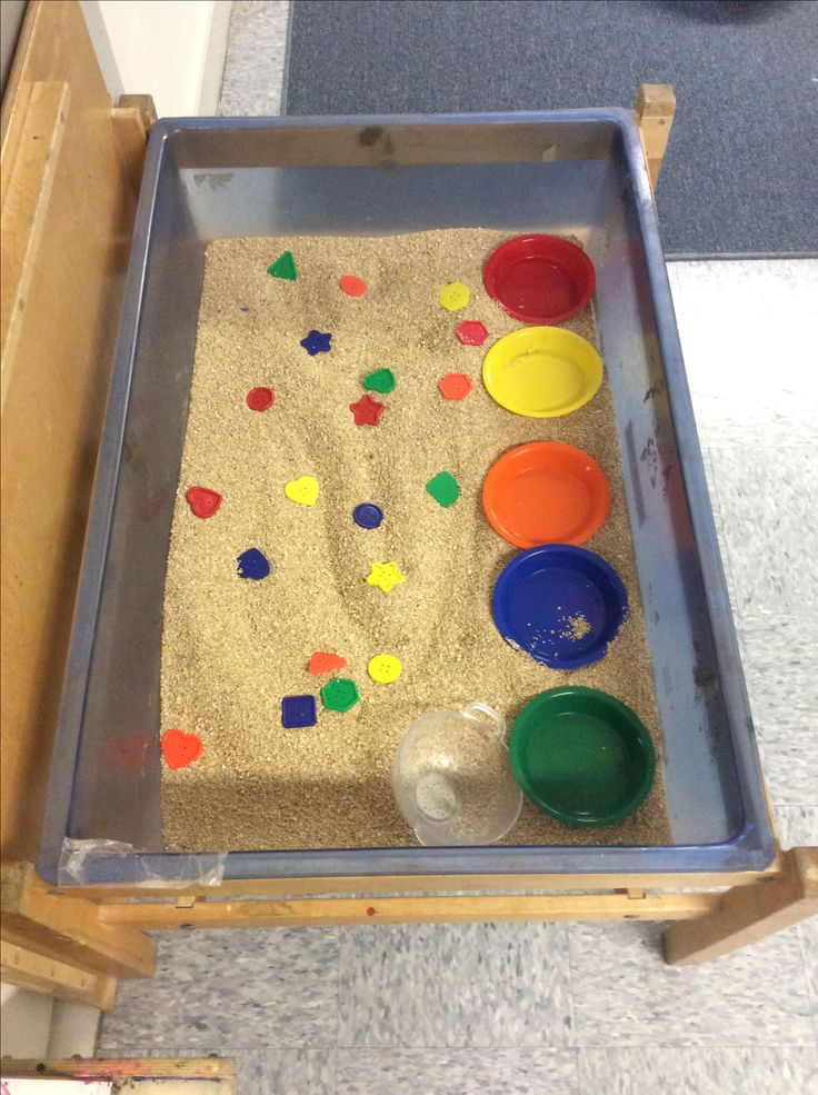 an empty tray with sand and paints on the floor next to a wooden box filled with plastic cups