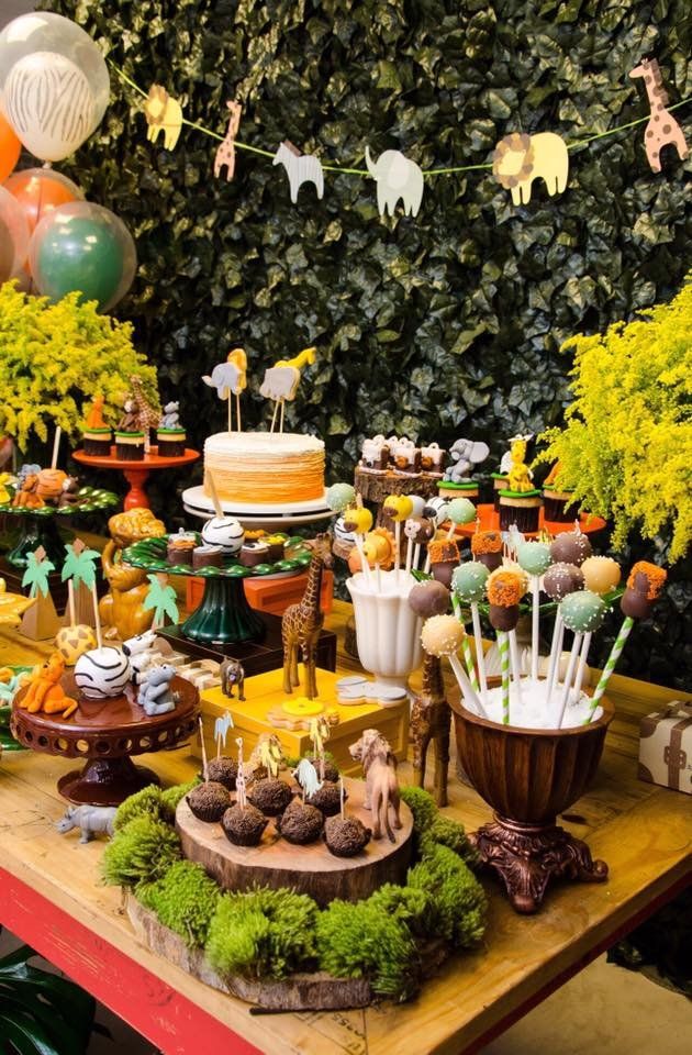 a table topped with lots of desserts next to green plants and balloons hanging from the ceiling