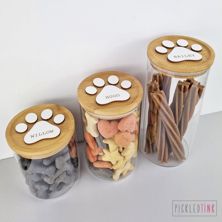 three jars filled with different types of dog treats and sticks in each jar, one has a paw print on the lid