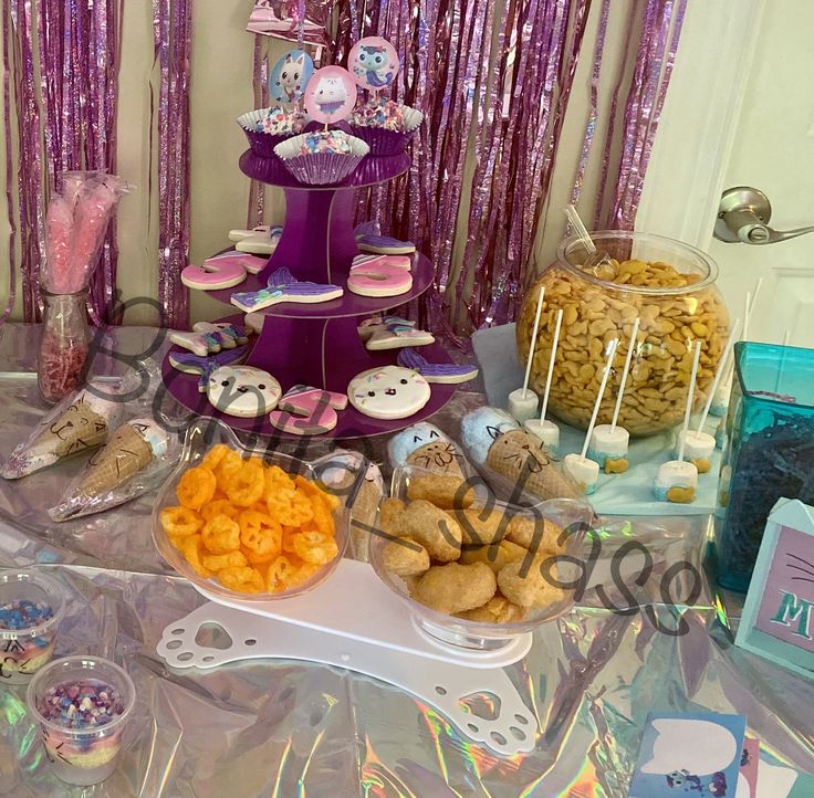 a table topped with lots of food and desserts next to purple foiled curtains