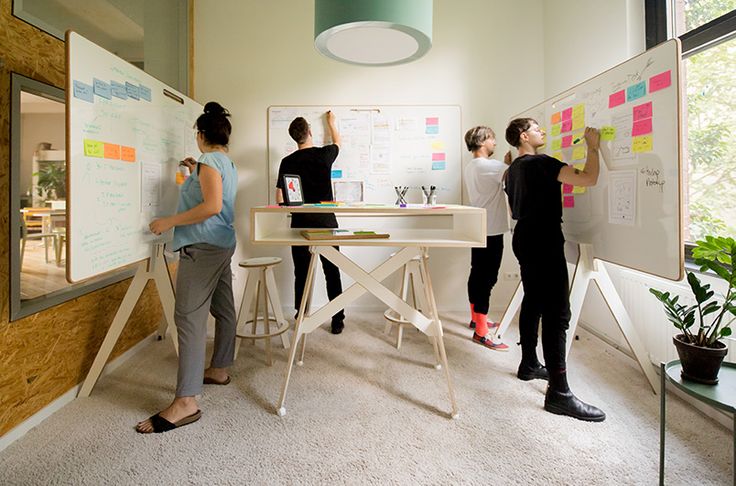 three women standing in front of a whiteboard with sticky notes on it and one woman pointing to the wall