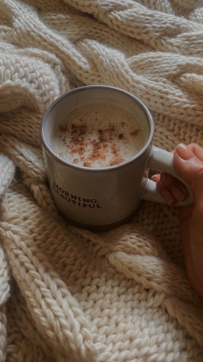 a person holding a cup of hot chocolate on top of a white knitted blanket