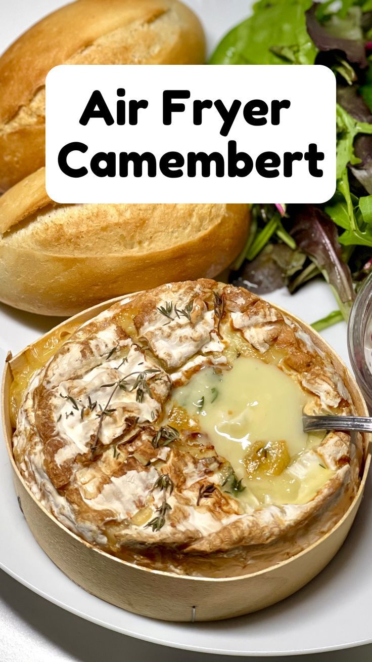 A whole camembert melting away on a white plate surrounded by bread rolls and salad, seen from above