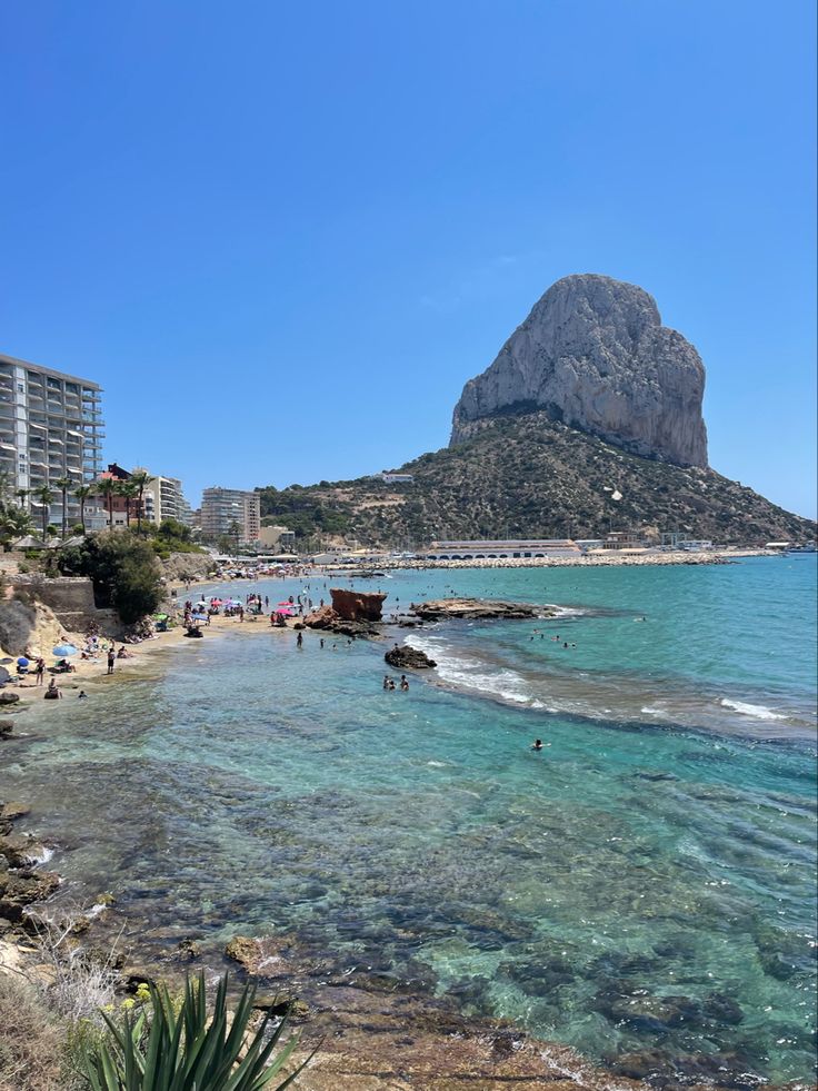 people are swimming in the clear blue water near an island with mountains and beach umbrellas