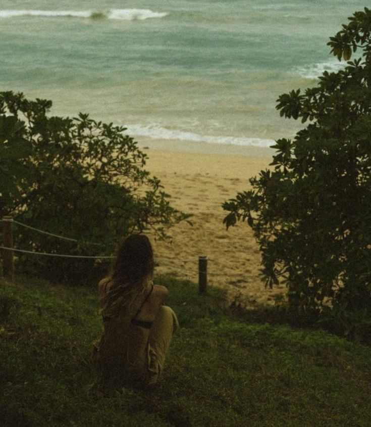 a woman sitting on top of a lush green hillside next to the ocean