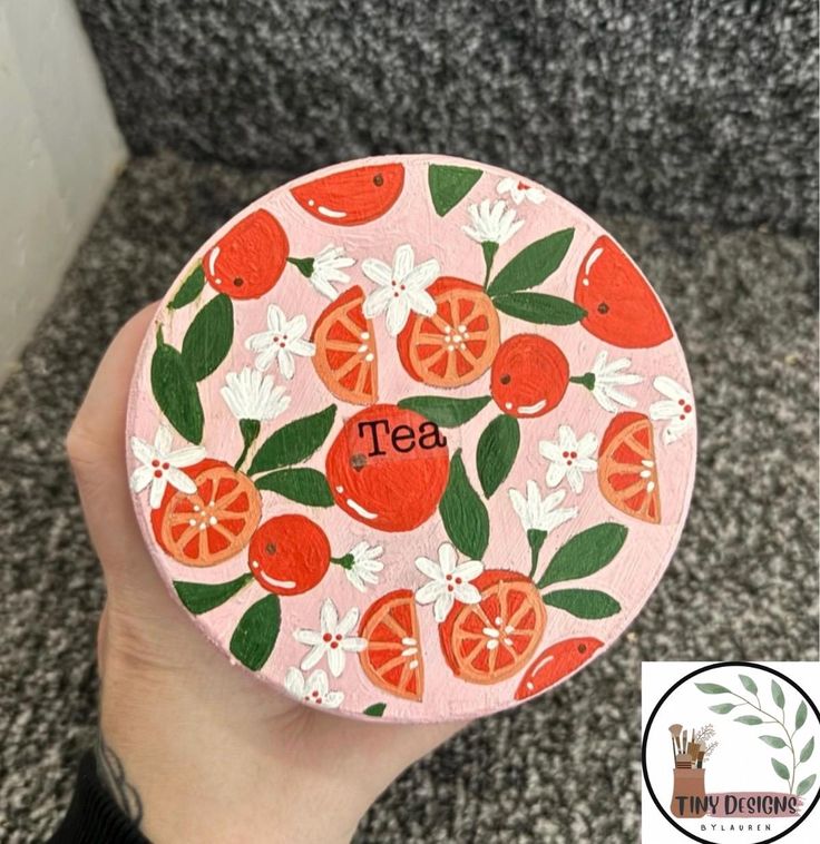 a hand holding up a pink coaster with oranges and flowers on it that says tea