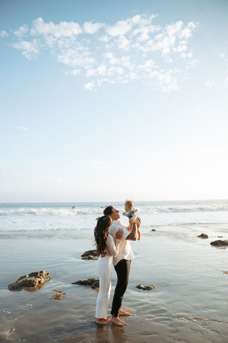 a woman holding a baby while standing on top of a beach next to the ocean