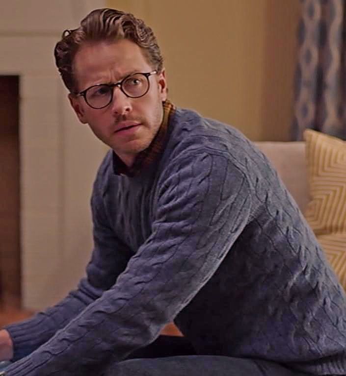 a man with glasses sitting on a couch in front of a fire place and looking at the camera