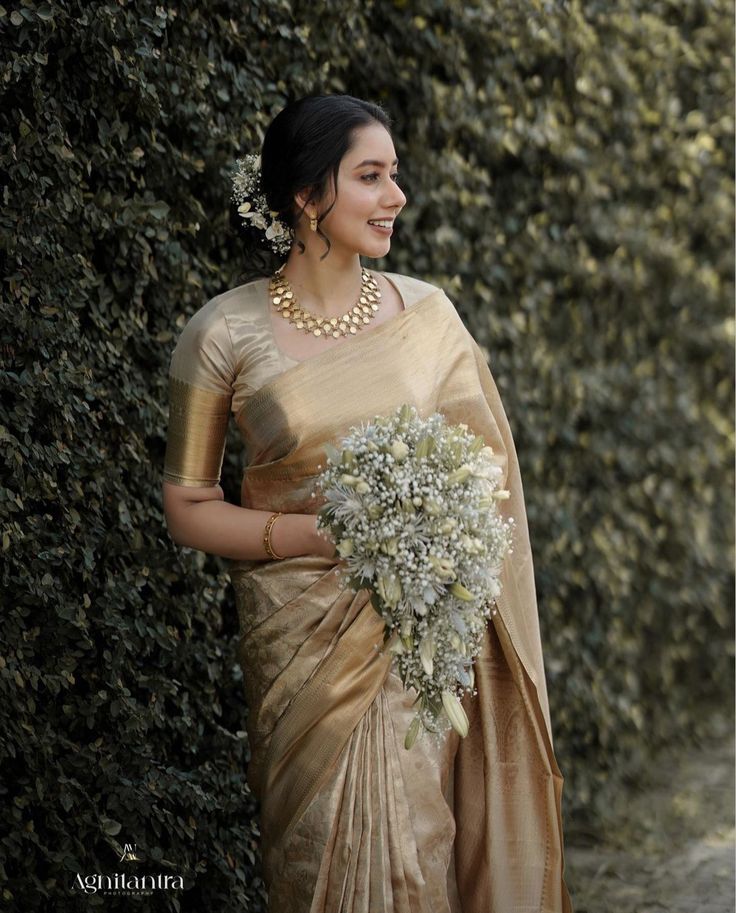 a woman in a gold sari holding a bouquet of white flowers and smiling for the camera