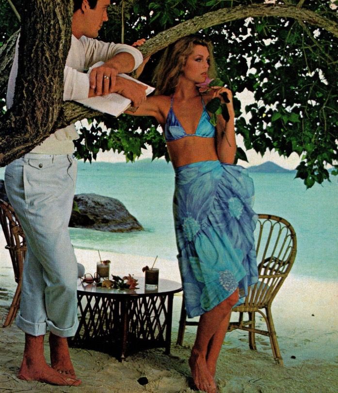 a man standing next to a woman under a tree on the beach with an umbrella