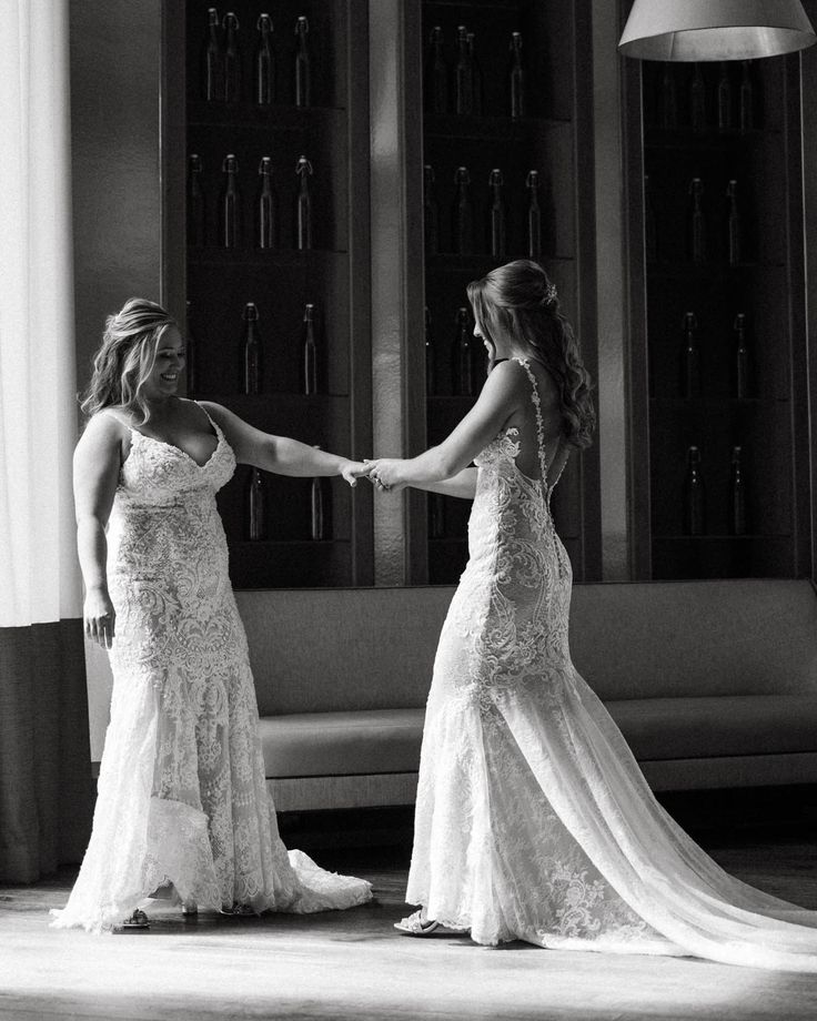 two women in wedding dresses holding hands and looking into each other's eyes while standing on the floor