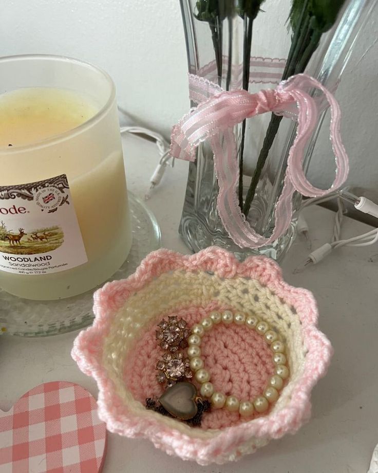 a pink crocheted bowl next to a candle and some flowers on a table