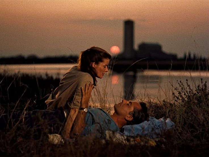 two people laying on the ground in front of water at sunset or sunrise, one person has his head up to the other's chest