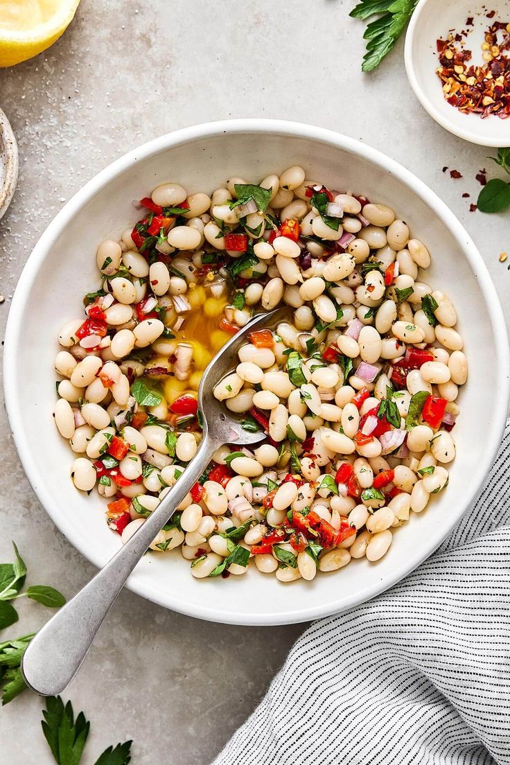 a white bowl filled with bean salad next to lemons and parsley on the side