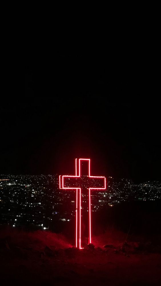 a lighted cross in the middle of a field at night with city lights behind it