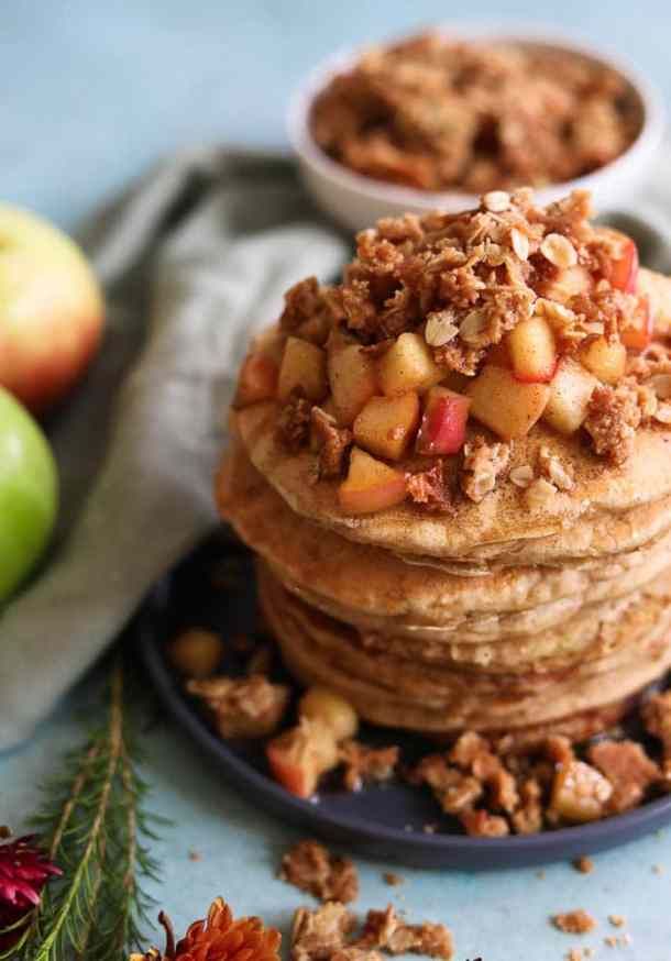 stack of pancakes with apples and granola toppings on blue plate next to flowers