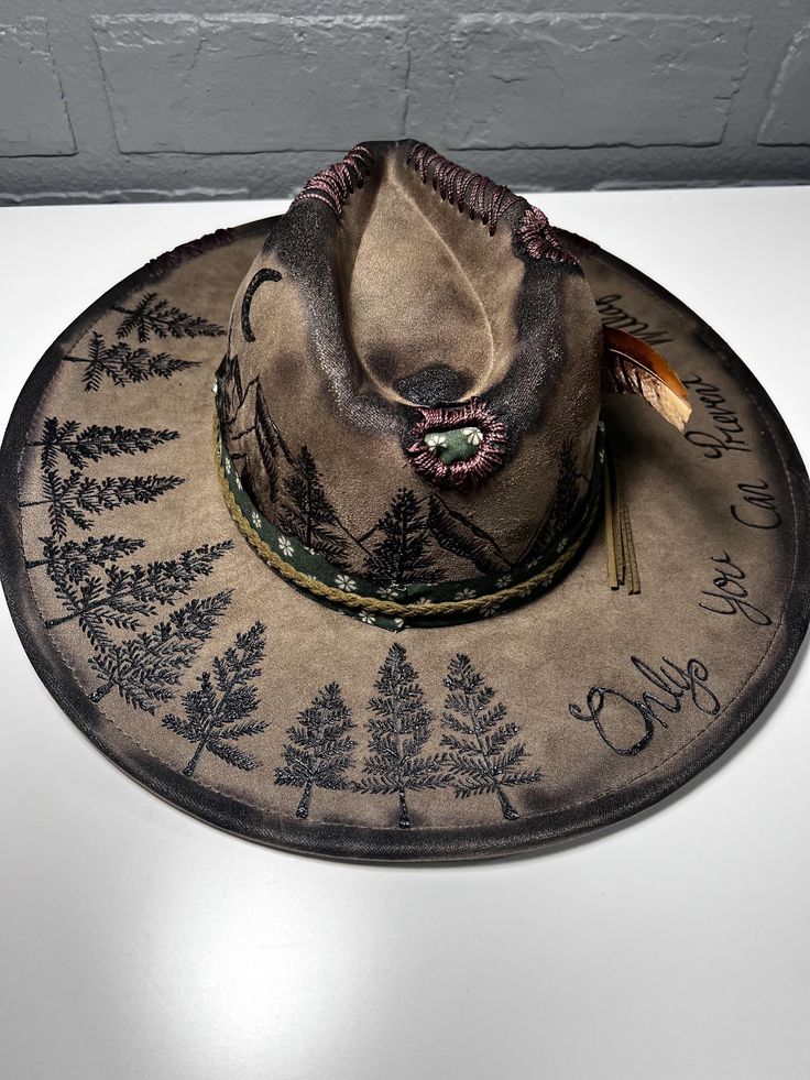 A hand-burned wide-brim hat with tree designs, mountain details, a feather accent, and "Only You can prevent forrest fires" written on the brim. Custom Burned Hat Ideas, Burned Designs On Hats, Rustic Curved Brim Hunting Hat, Rustic Brimmed Felt Hat For Outdoor, Rustic Outdoor Brimmed Felt Hat, Rustic Flat Brim Felt Hat For Outdoor, Rustic Felt Hat With Flat Brim For Outdoor, Handmade Country Hat For Outdoor, Handmade Country Style Outdoor Hat