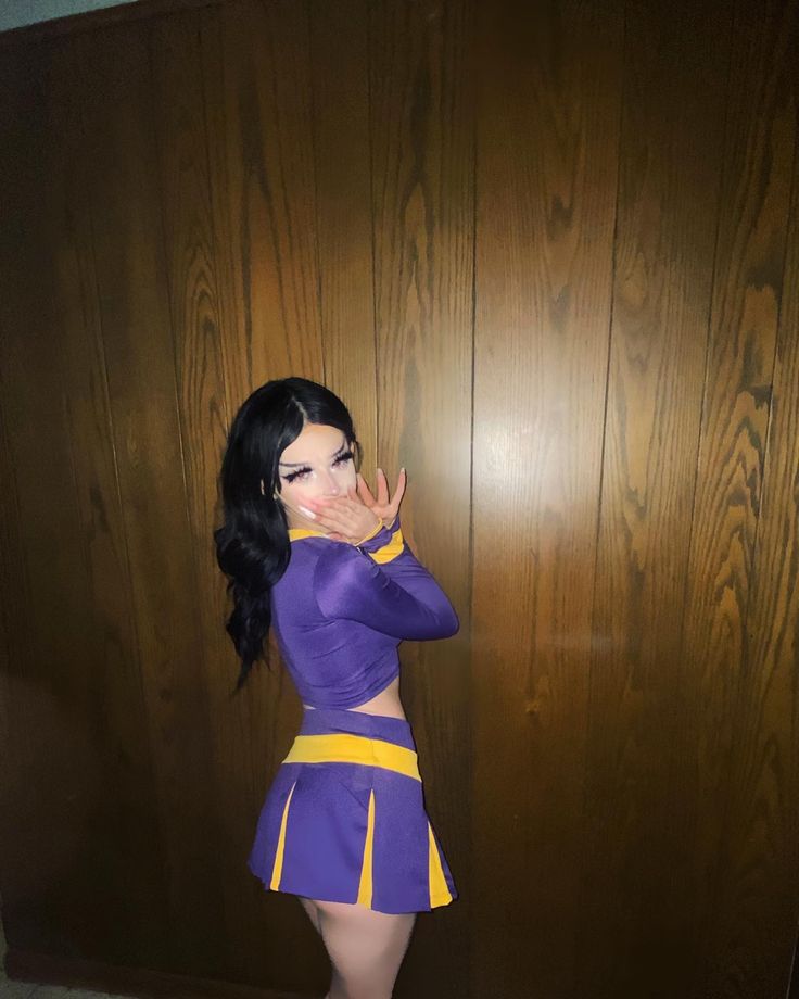 a woman dressed in a purple and yellow outfit standing next to a wooden wall with her hand on her face