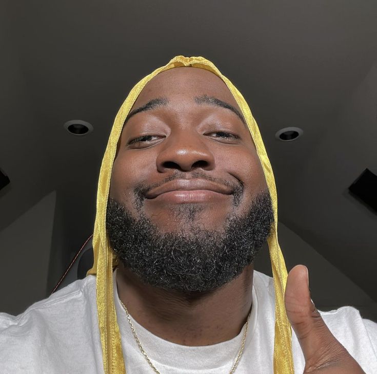 a man with a beard and yellow scarf around his head smiles at the camera while wearing a white t - shirt