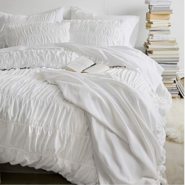 a bed with white comforter and pillows in a room next to a stack of books