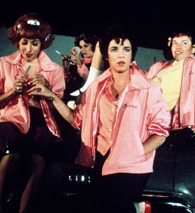 three women in pink shirts and black pants are sitting on a bench with one woman cutting another's hair