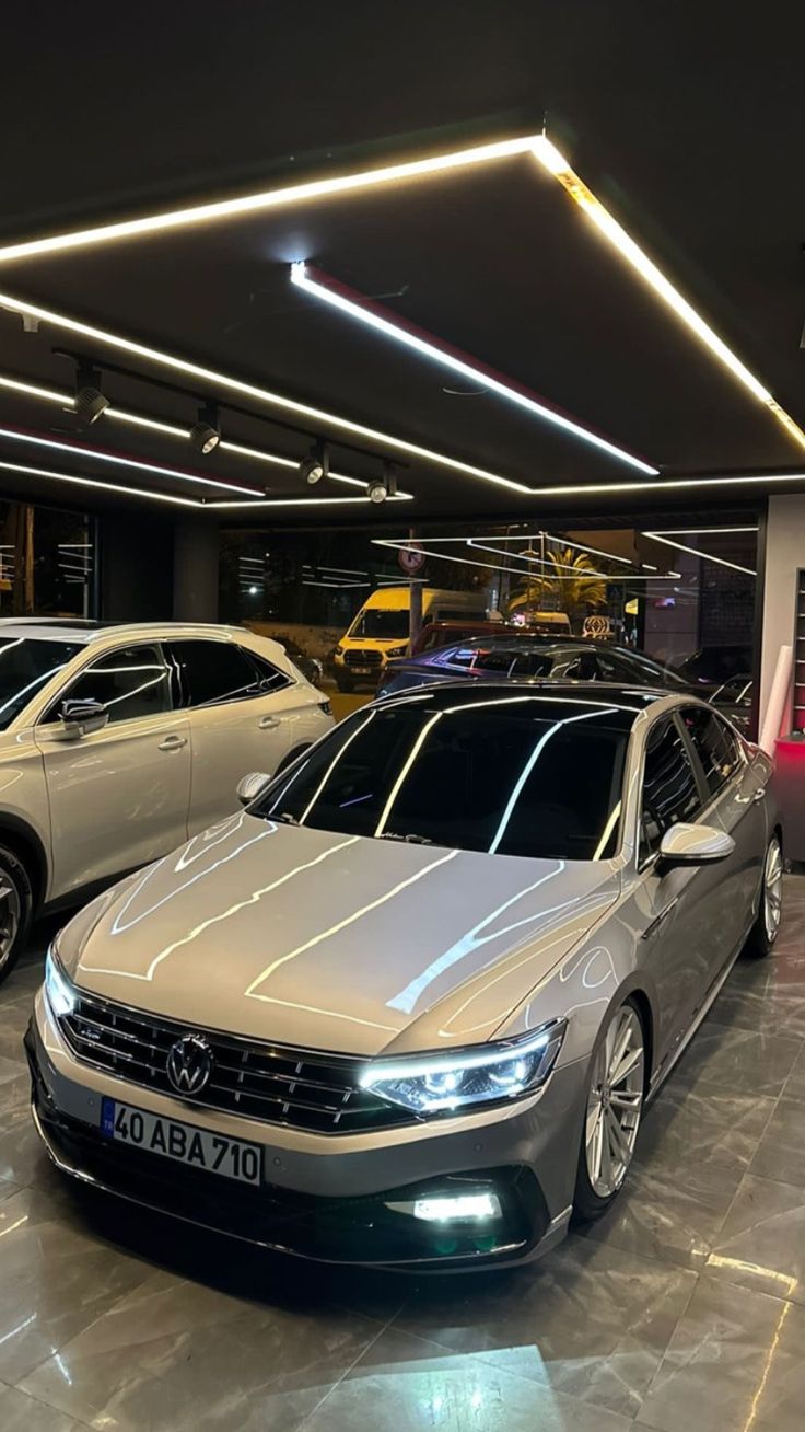 two cars are parked in a garage with lights on the ceiling and below them is a car showroom