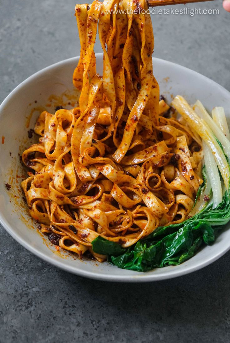 someone is holding chopsticks above a bowl of noodles and vegetables on the table