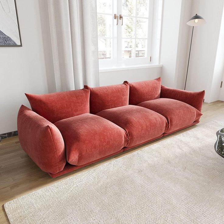 a red couch sitting on top of a wooden floor next to a white rug in front of a window