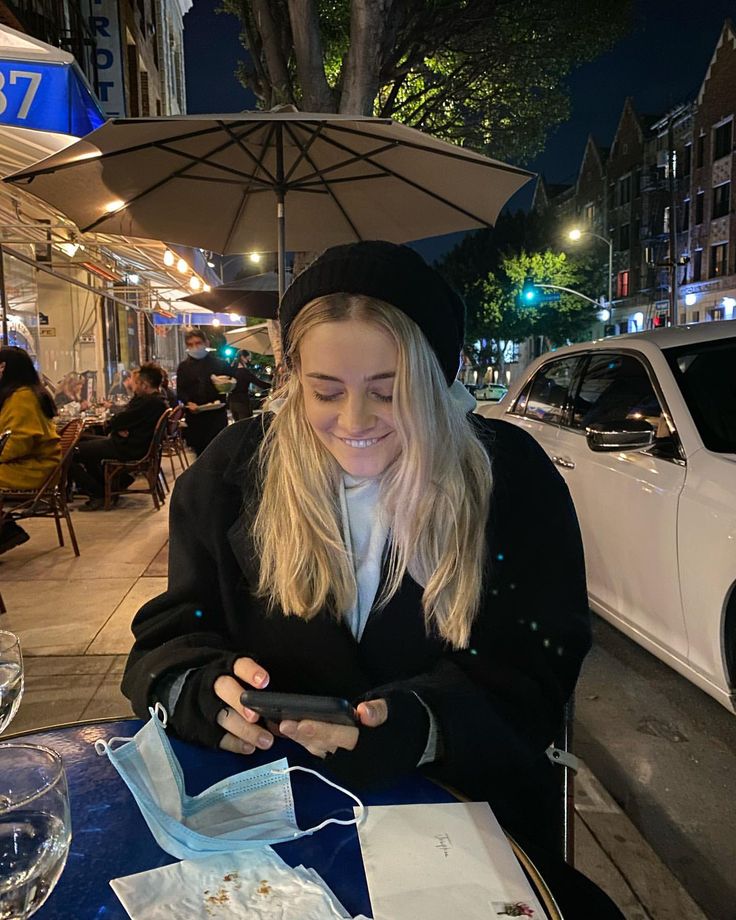 a woman sitting at an outdoor table looking at her cell phone while wearing a beanie