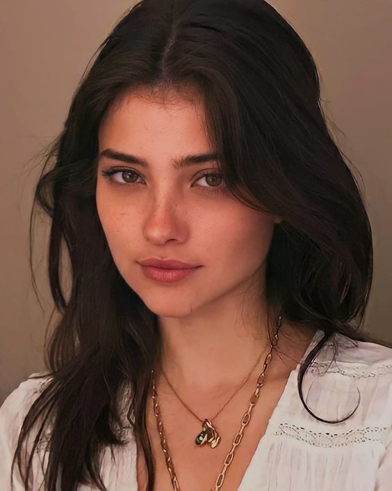 a woman with long hair wearing a white shirt and gold necklace, looking at the camera