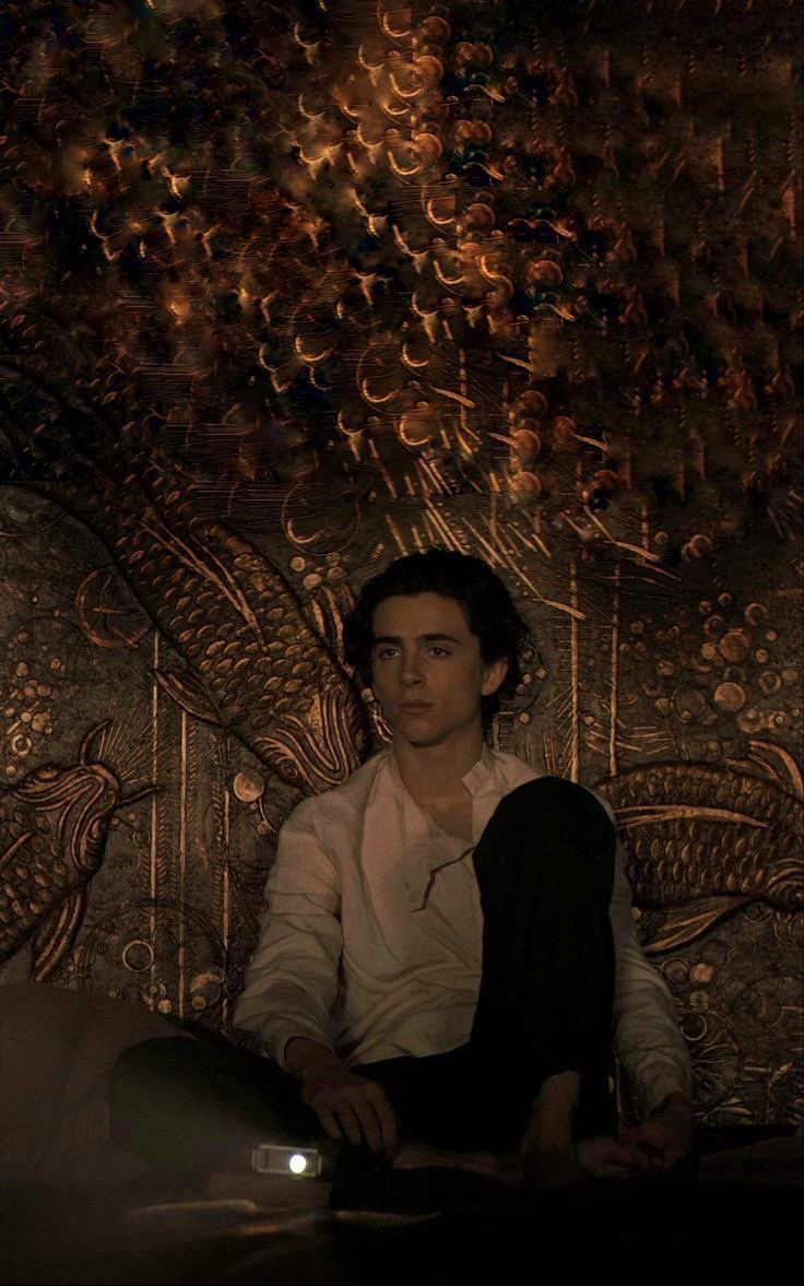 a young man sitting on top of a bed in front of a wall covered with intricate designs