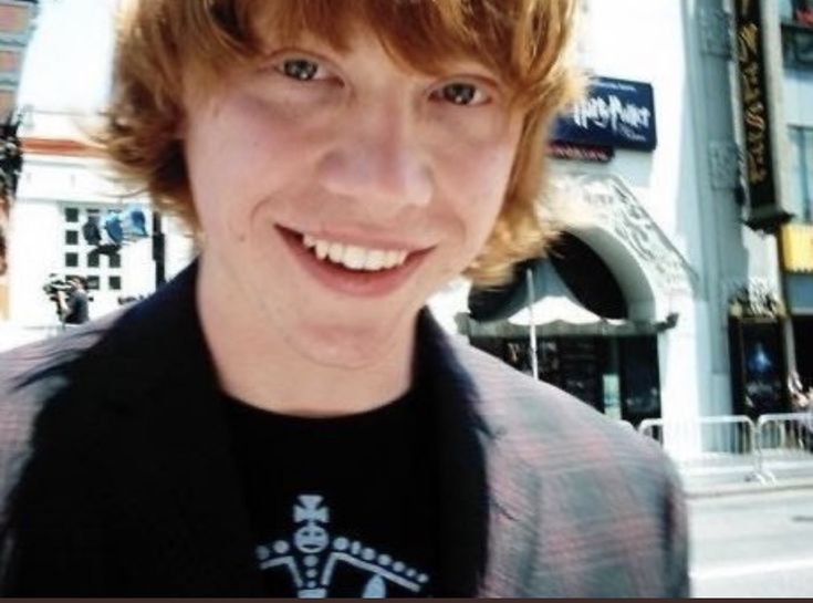 a young man with red hair and blue eyes smiles in front of a city street