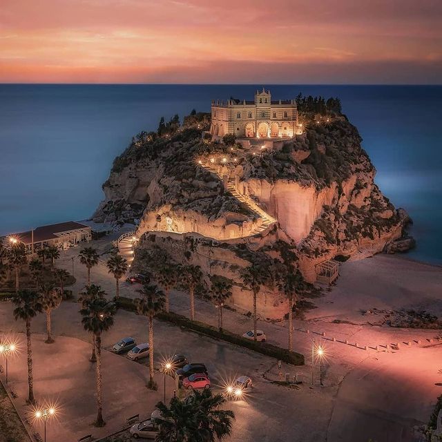 a castle on top of a hill next to the ocean at night with lights lit up