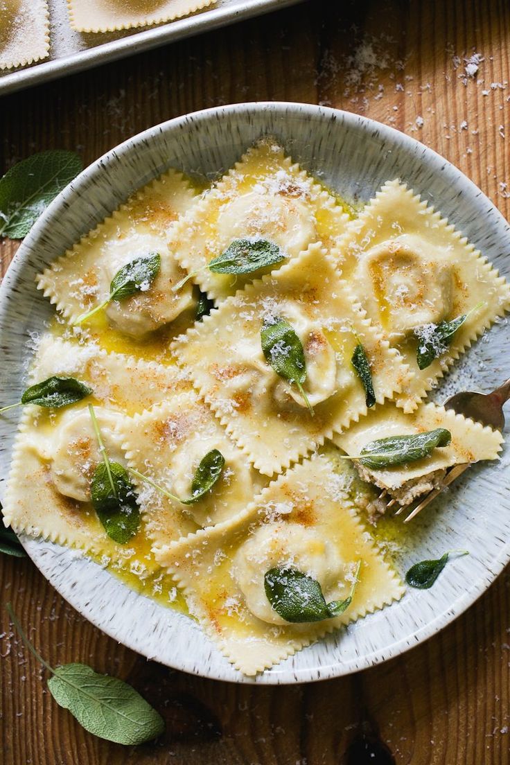 a white bowl filled with ravioli and sauce on top of a wooden table next to green leaves