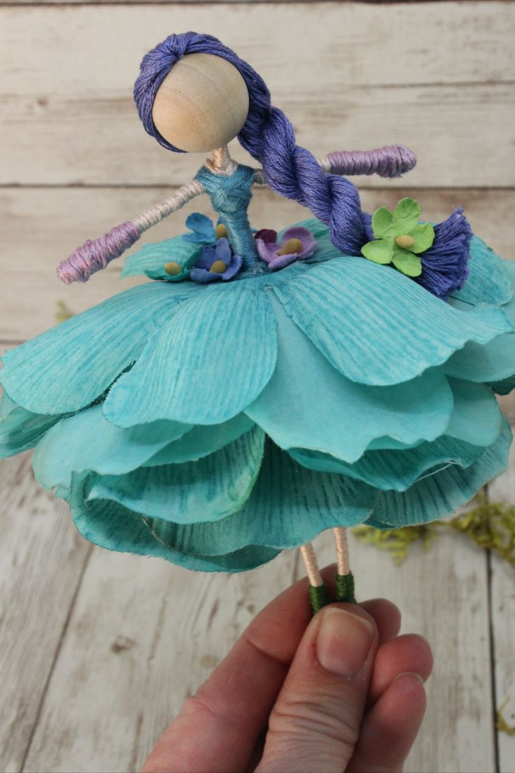 a hand holding up a doll made out of blue paper and green yarn with flowers on it