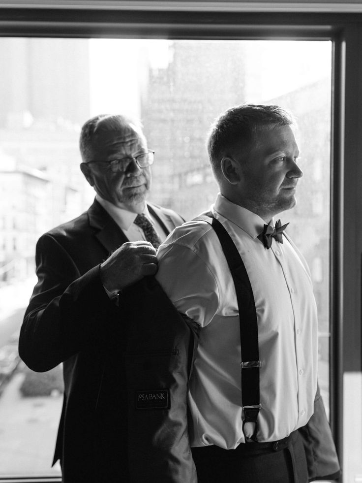 two men standing next to each other in front of a window with one adjusting the tie