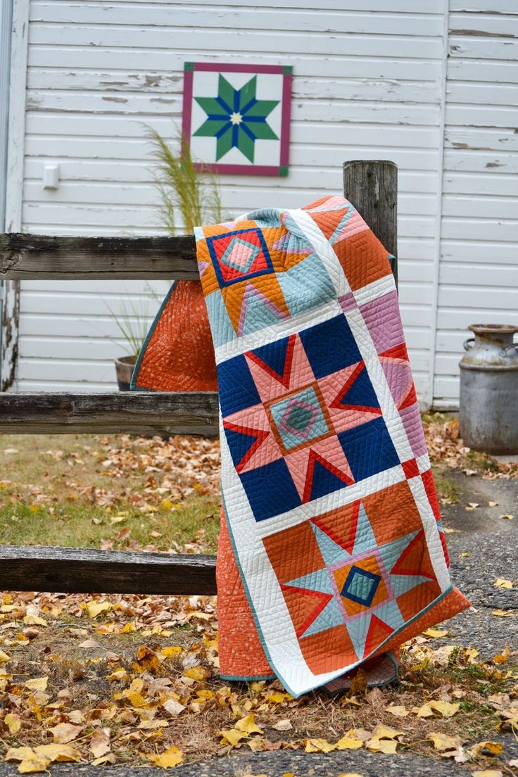 an orange and blue quilt sitting on the ground next to a white building with a wooden fence