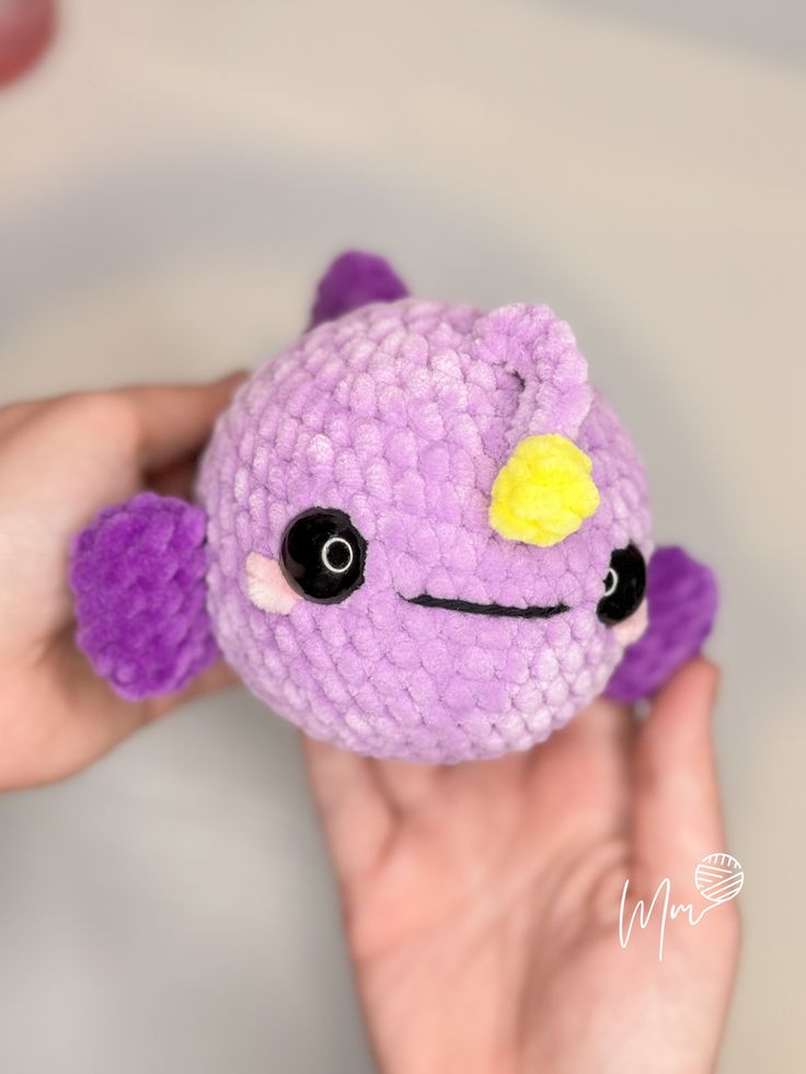 a hand holding a purple stuffed animal with black eyes and yellow eye patch on its nose