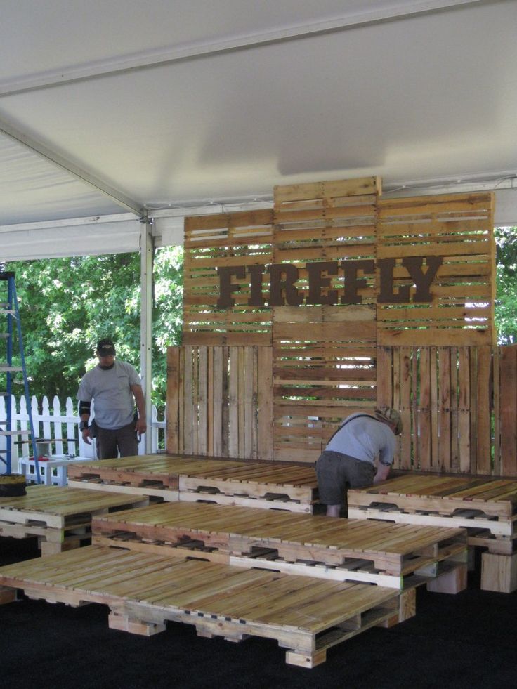 two men working on some wooden pallets under a tent with trees in the background