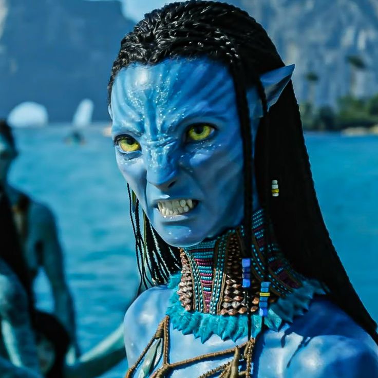 a woman with blue makeup and yellow eyes standing in front of the ocean while other people watch