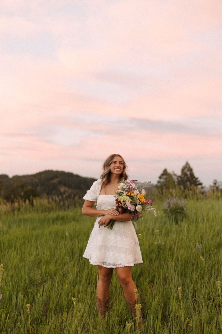 a woman in a white dress is standing in tall grass holding flowers and looking up at the sky