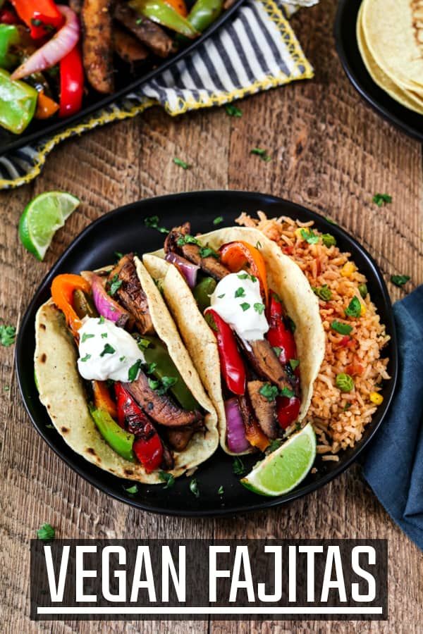 two tacos with meat, peppers, onions and rice on a black plate next to lime wedges