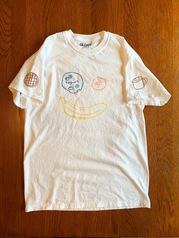 Brunch T-Shirt Hand drawn with fabric marker on white short sleeve unisex Gildan t-shirt, 100% cotton Features designs on front (blueberry yogurt, cutie, banana), left sleeve (stroopwafel), right sleeve (coffee cup) and back (artist's name) Small and Medium size available Note: Wash inside out on cold. Marker may fade a little after initial wash. No worries, it still looks great! Each shirt will either be shipped via bubble mailer or tube. Design by Josh Bokor White Short Sleeve T-shirt With Funny Print, Pre-shrunk Organic Cotton Short Sleeve Shirt, Unisex White T-shirt With Funny Print, White Cotton Shirt With Cartoon Print, Unisex White T-shirt With Cartoon Print, White Short Sleeve T-shirt With Cartoon Print, Cute Short Sleeve T-shirt With Screen Print, Short Sleeve T-shirt With Funny Print, White T-shirt With Funny Print, Relaxed Fit