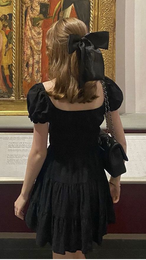 a woman standing in front of a painting wearing a black dress with a large bow